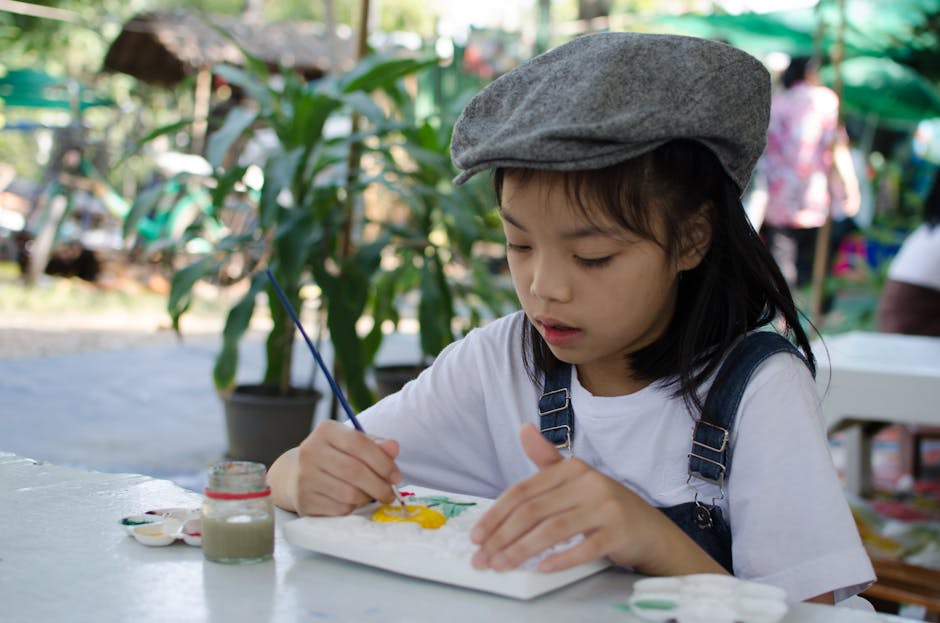A young girl in a hat painting with watercolors on a sunny day outdoors.